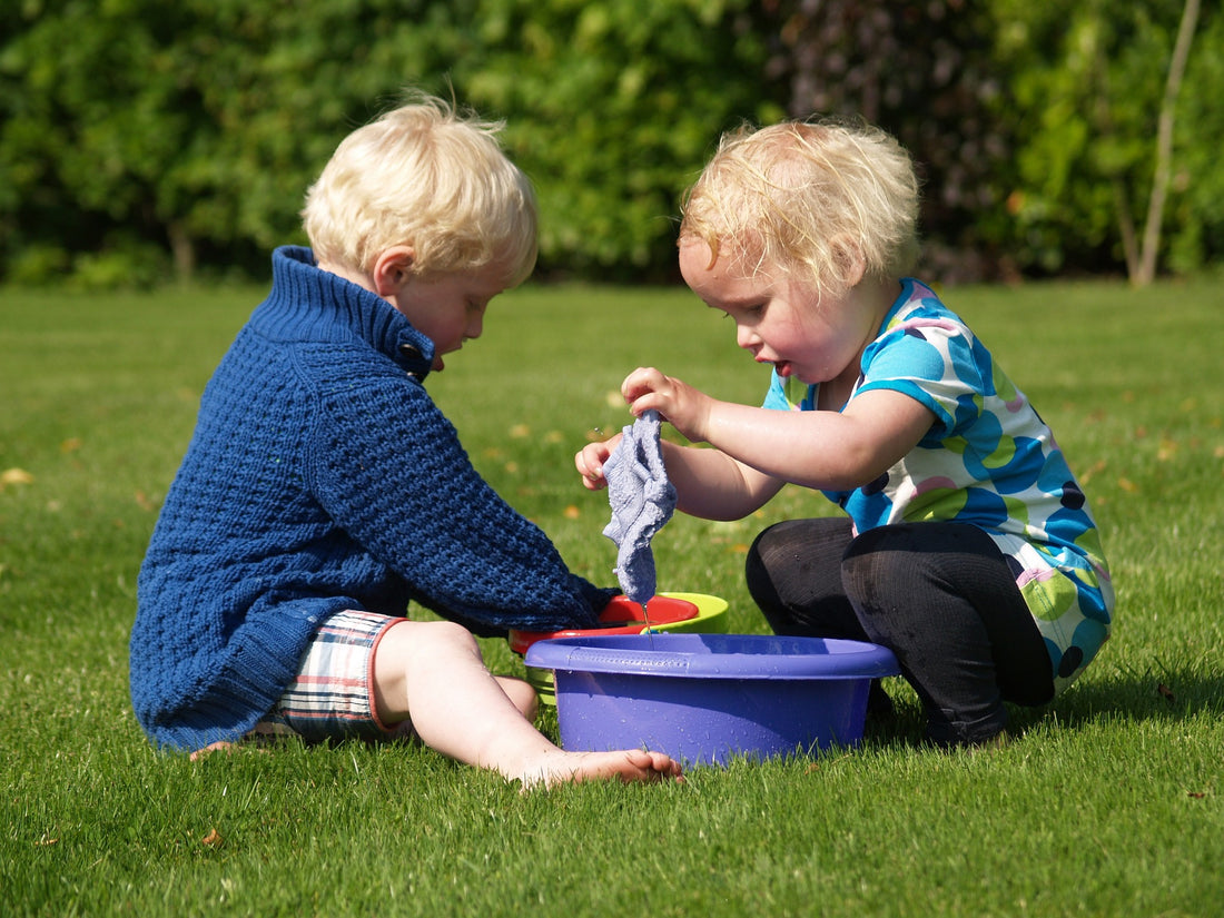 Two toddlers play with a washcloth and container of water outside. Two-year-olds are at the stage of development in which they are engaging in symbolic and sequence play.