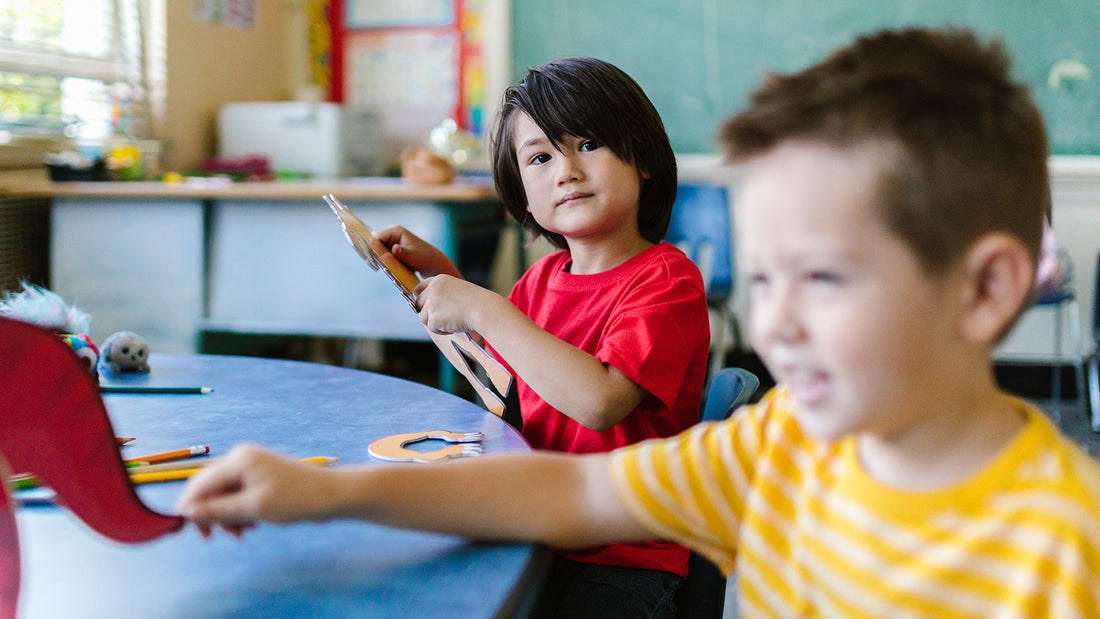Two young boys sit at a table at school, looking at the camera while holding pieces of a puzzle
