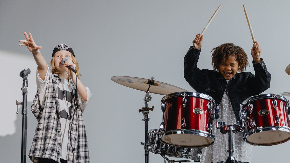 A young girl laughs and plays the drums as her friend sings into a microphone