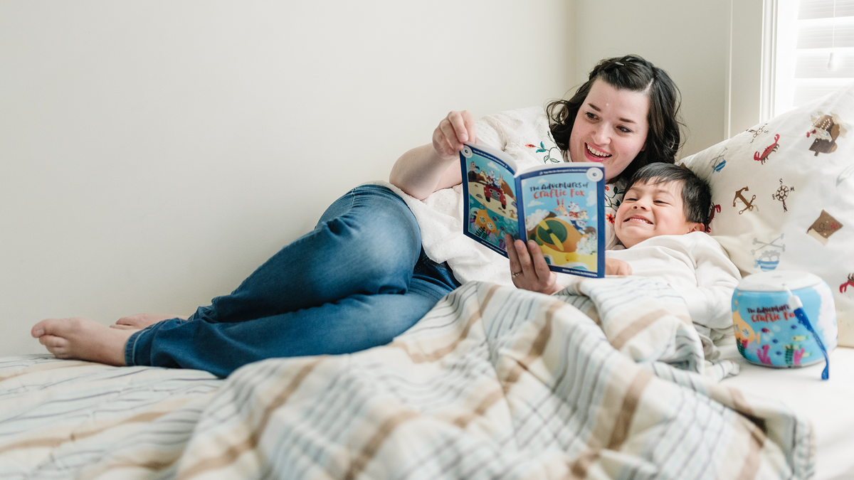 Mother and child cuddle in bed while reading "The Adventures of Craftie Fox" with a Storypod nearby.