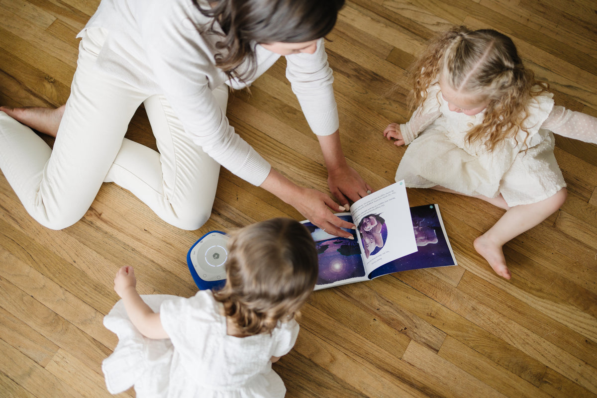 Mother sits on the floor with two young daughters, flipping through a book while using Storypod. Engaging in audio experiences is one way to make reading fun for the reluctant child.