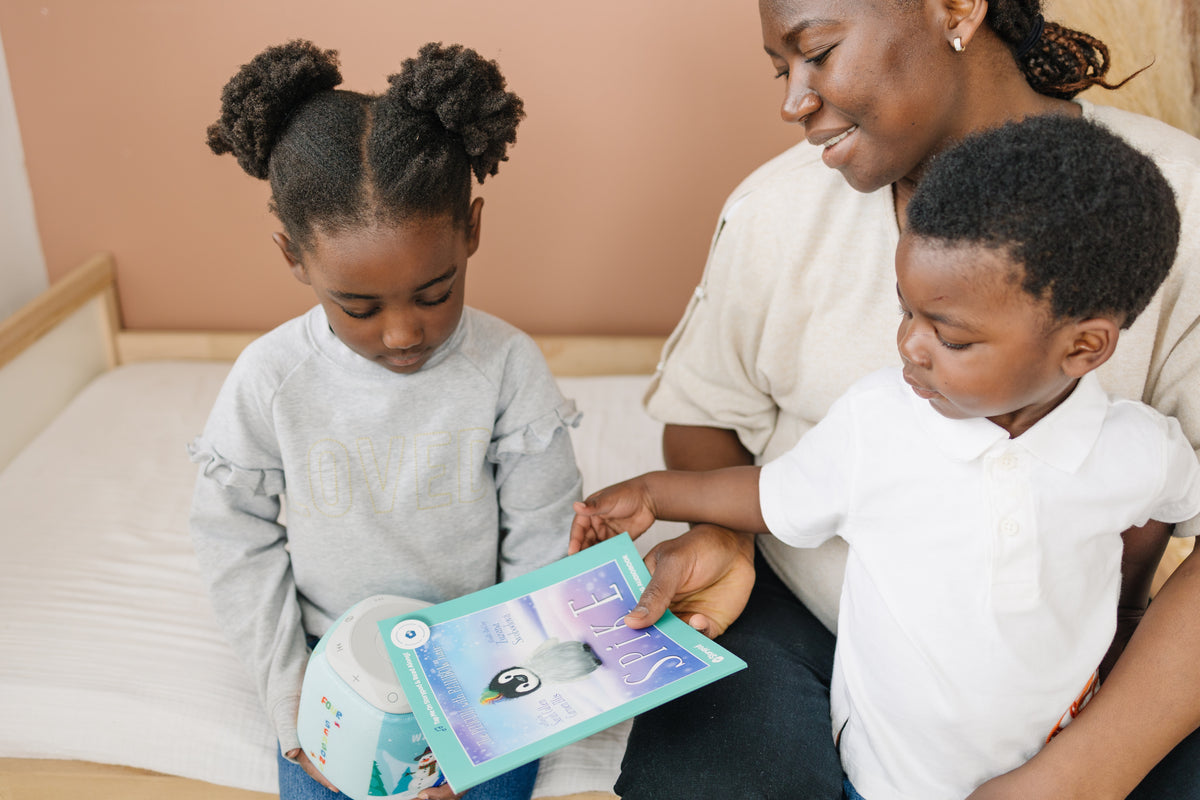 Mother sits on a bed with a toddler on her lap and her 4-year-old daughter by her side. Practicing reading comprehension skills even before your child can read will give benefit your child greatly.