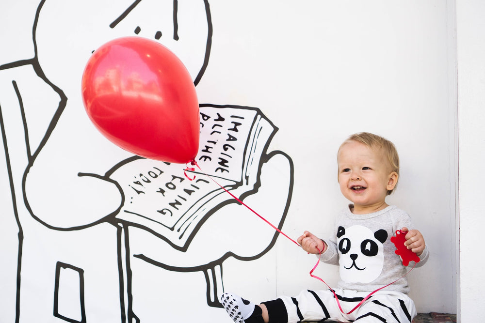 One-year-old child sits in front of a simple mural while holding a bright, red, birthday balloon. A parent can look forward to many strides in their child's development at one-year-old.