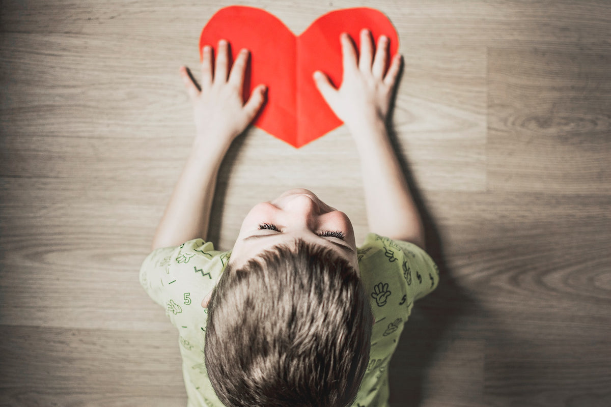 Little boy lies on the floor with a red, paper heart in his hands. Teaching your child to perform simple acts of kindness is a great way to show love in February.