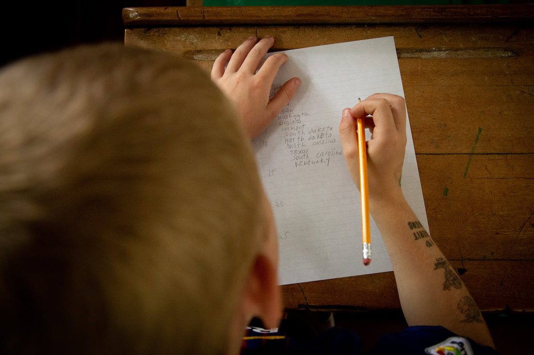 A first grade boy sits at a desk writing on a piece of paper. Providing your little one with creative story-starters is a great way to encourage them to practice their writing skills.