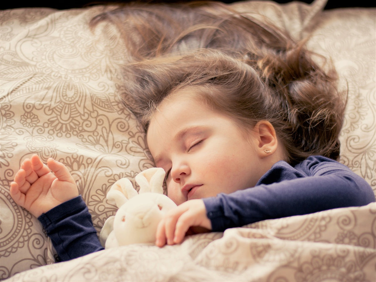 1-year-old girl sleeps in bed accompanied by her stuffed bunny. It's important to develop a healthy sleep routine for your child so they can reap the maximum health benefits.