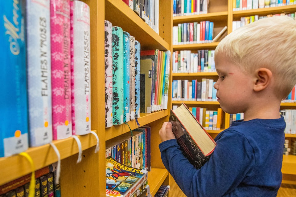 2-year-old boy looks through books at the library. Although your toddler isn't able to read on their own yet, there are several pre-literacy skills you can start building on now.