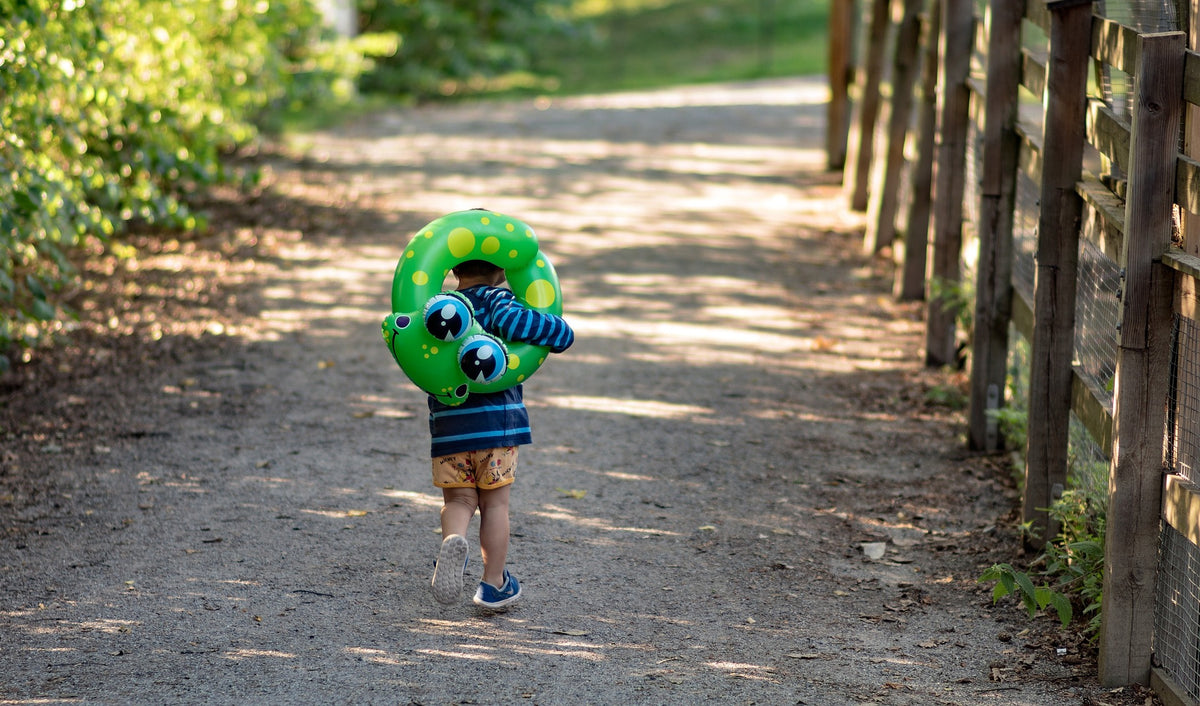 2-year-old child walks down a path while holding an inflatable from-themed pool floatie. Gross motor skills refer to the physical abilities that involve the use of large muscles in the body and are rapidly developing in your child at this age.