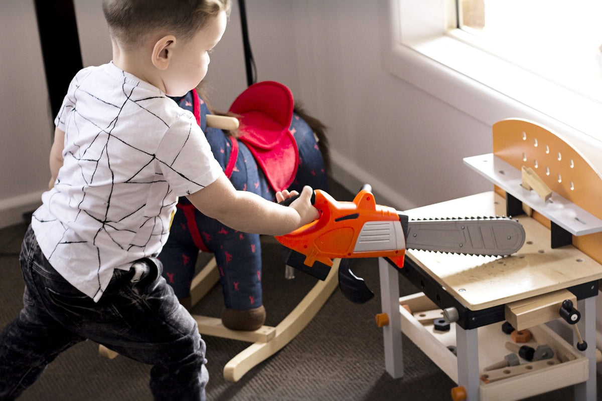 1-year-old boy plays with a toy saw at a miniature, wooden work bench. Toys that emulate what children see adults doing are bound to hold their attention.
