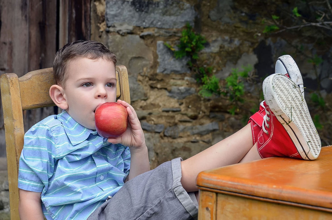 5-year-old boy sits with his feet on the desk and eats an apple. Teaching your little one healthy habits, like eating the rainbow, is important for their well-being.