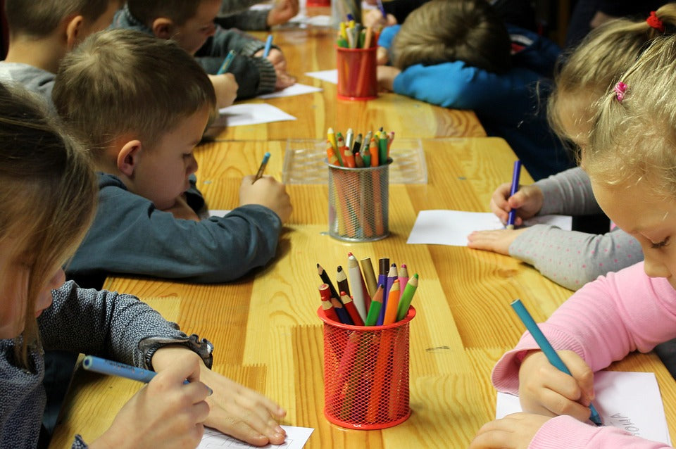 1st grade students sit a table working using colored pencils and paper. In the 1st grade, your child will be learning to write simple sentences accompanied by a drawing to express their ideas.