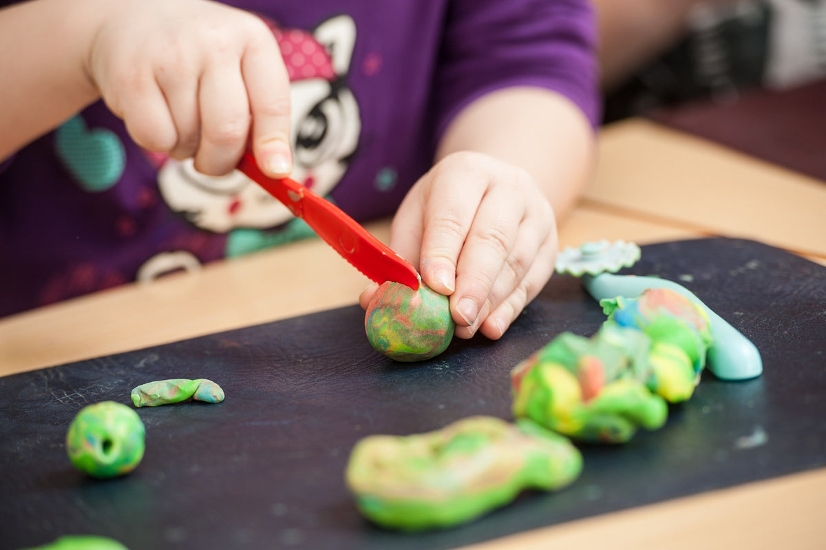 A pair of child's hands cut a ball of play dough with a toy knife. Using pray dough to recreate sight words is one fun way to practice this important literacy skill.