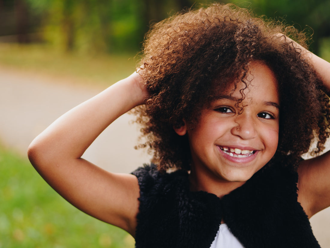 Six-year-old girl flashes a big smile while playing with her curly hair. A child's 6th year is marked by an increase in independence and rapid development of mental skills.