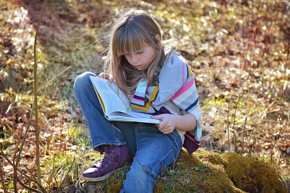 6-year-old girl sits on a rock outside while reading a book. Motivating your child to read is important for their literacy development.