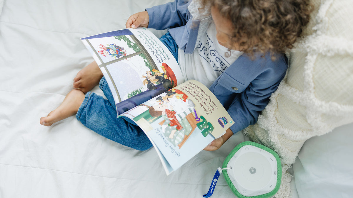 Toddler sits on bed with book on his lap and a Storypod beside him