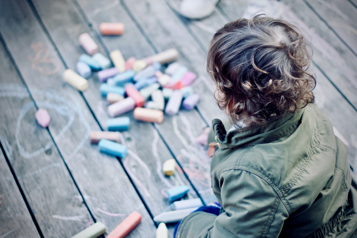 Child sits on a deck drawing with sidewalk chalk. Using fun materials is a great way to incentivize your child to practice writing.