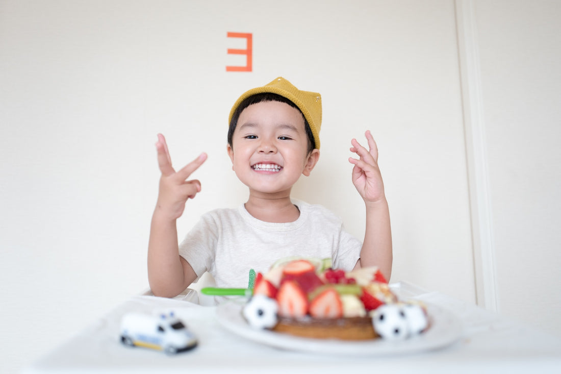 3 year old boy in yellow hat holds up 3 fingers in front of his birthday cake. There are many milestones to look forward to in your child's 3rd year.