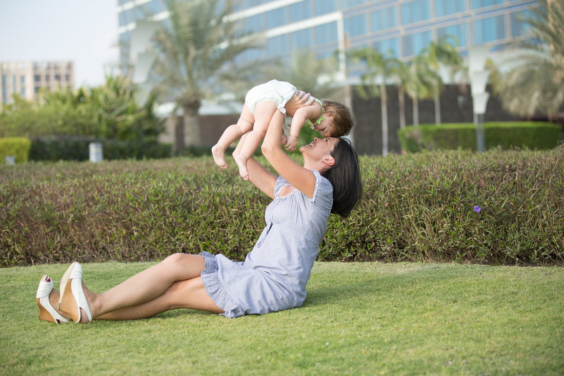 Mother gleefully lifts her 1-year-old daughter in the air while sitting outside. 1-year-olds can understand many things, including emotions and several words and phrases.