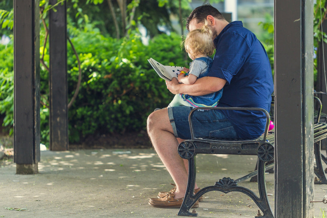 A father holds his 1-year-old daughter in his lap while reading her a book on an outside bench. Although 1-year-olds may not understand everything you are reading to them, they are gaining a lot from having a regular reading routine.