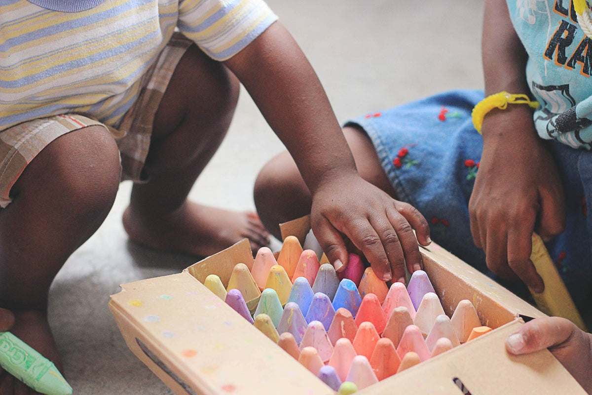 2-year-old boy squats down to a box of colorful sidewalk chalk. Engaging your child in low-mess art projects are a great way to bond with them while practicing motor skills and exercising their creativity.