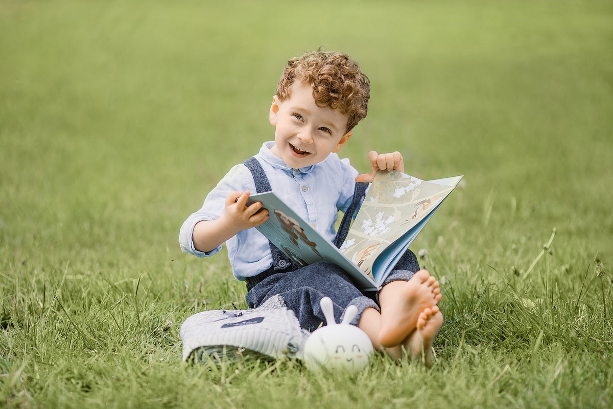 4-year-old boy sits in a grass field while reading a book. Reading books is one of the best ways to grow your child's descriptive vocabulary.