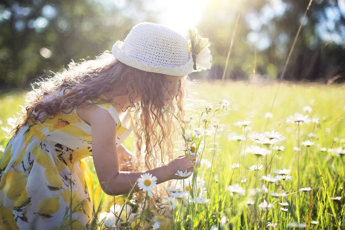 5-year-old girl in a yellow dress and white hat bends over to pick some white flowers. Creating nature centered crafts is a great way to encourage your child to spend time outdoors while fostering their creativity.